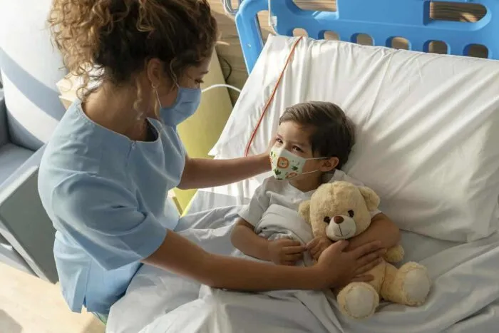 Boy lying down on hospital bed and sweet nurse comforting him both wearing facemasks