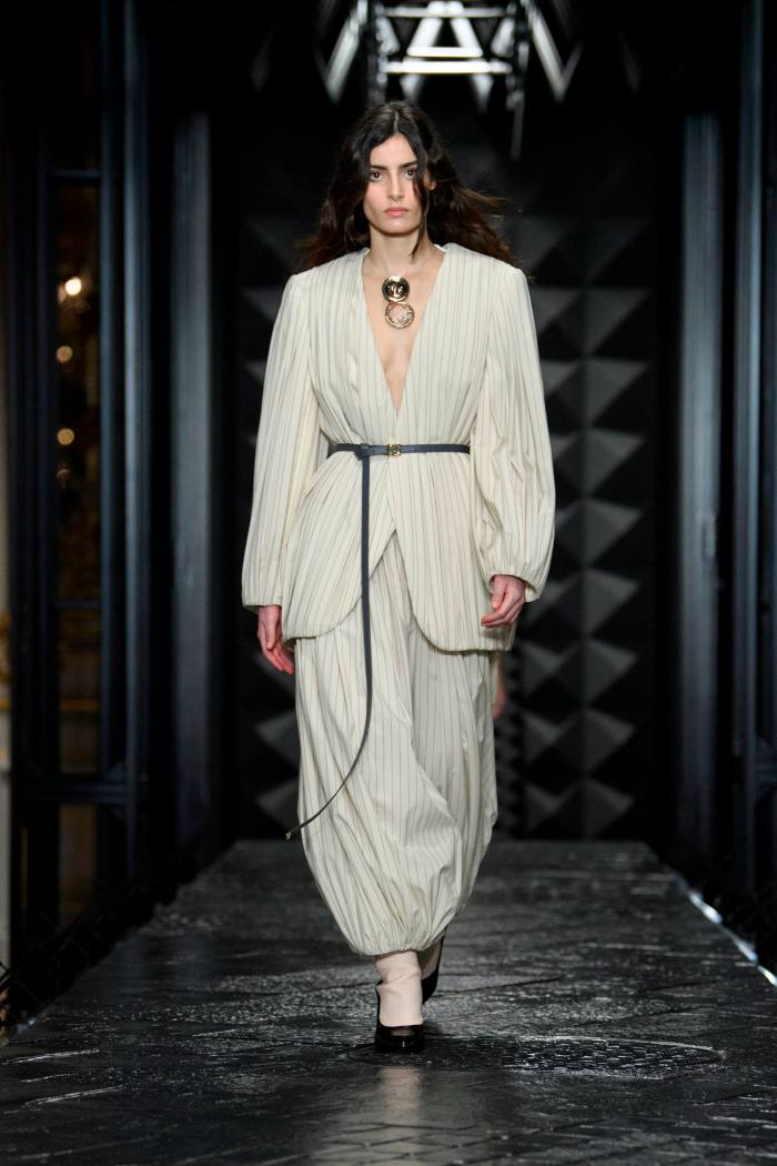 Un model feminin de podium într-o combinație lejeră de bluză deschisă și pantaloni din material mătăsos de culoarea fildeșului
