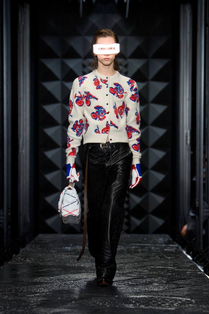Un model feminin de podium într-un cardigan alb cu model, pantaloni negri — și mască de față albă