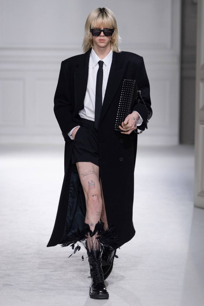 Un model feminin de podium în ochelari întunecați, blazer și cravată neagră, haină lungă neagră – și fustă scurtă cu cizme înalte