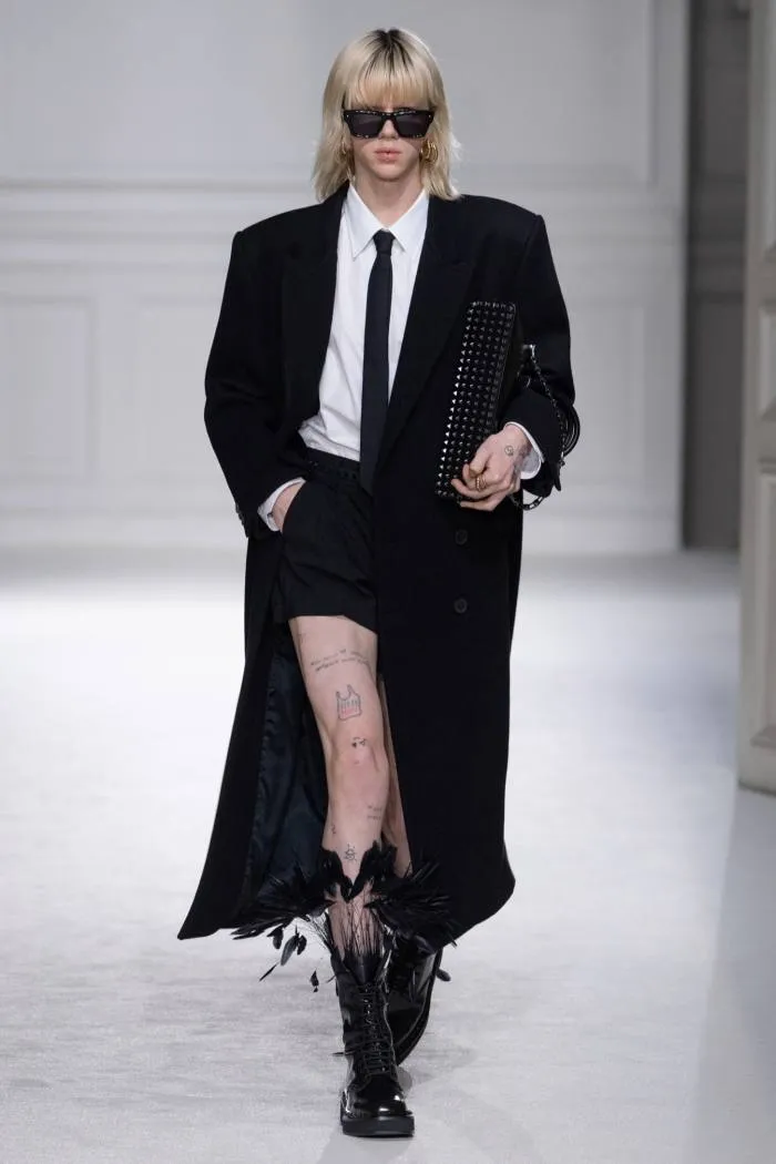 Un model feminin de podium în ochelari întunecați, blazer și cravată neagră, haină lungă neagră – și fustă scurtă cu cizme înalte