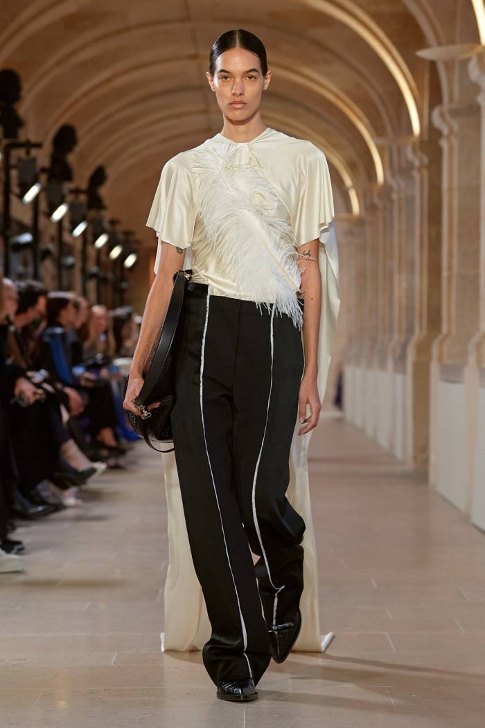 Un model androgin de podium în pantaloni scurți mătăsos, împodobiți cu un decor în formă de pene și pantaloni voluminosi mătăsos negri