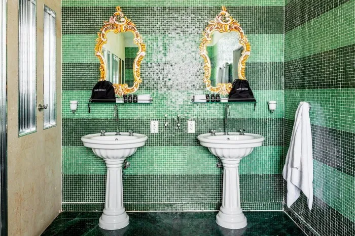 Două chiuvete albe și două oglinzi cu rame aurite într-o baie cu gresie verde