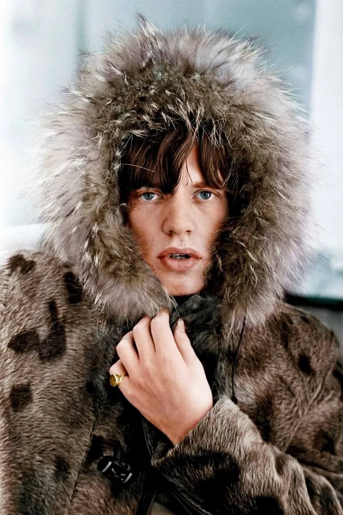 Mick Jagger se uită la cameră, purtând o haină de blană cu gluga trasă în jurul feței