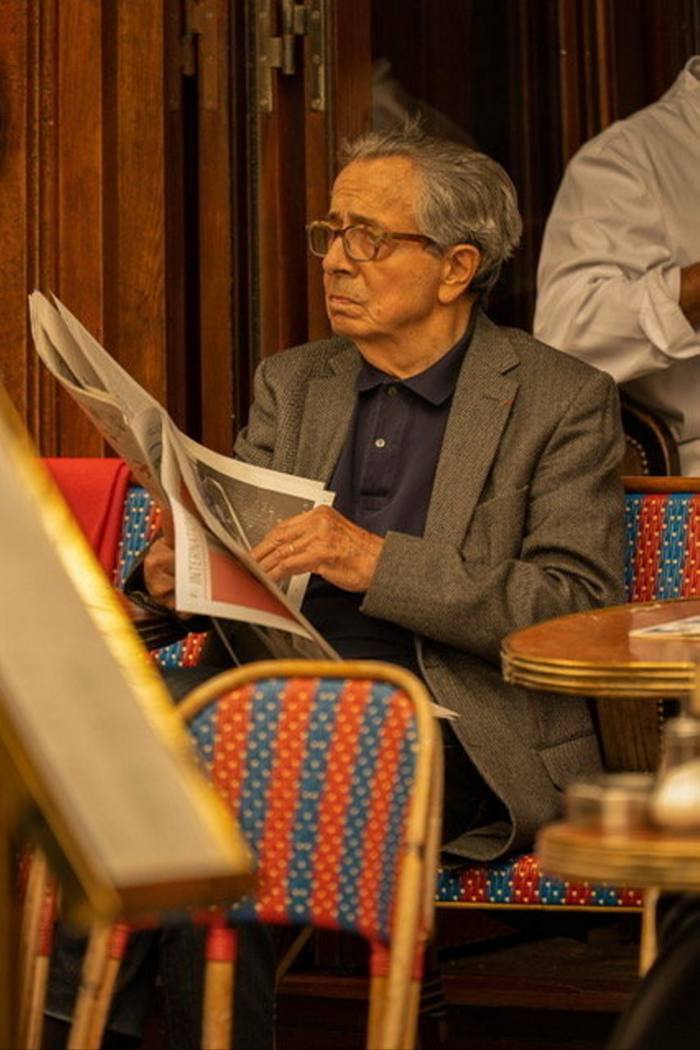 Un bătrân într-o jachetă gri, cămașă neagră într-o cafenea, citind un ziar