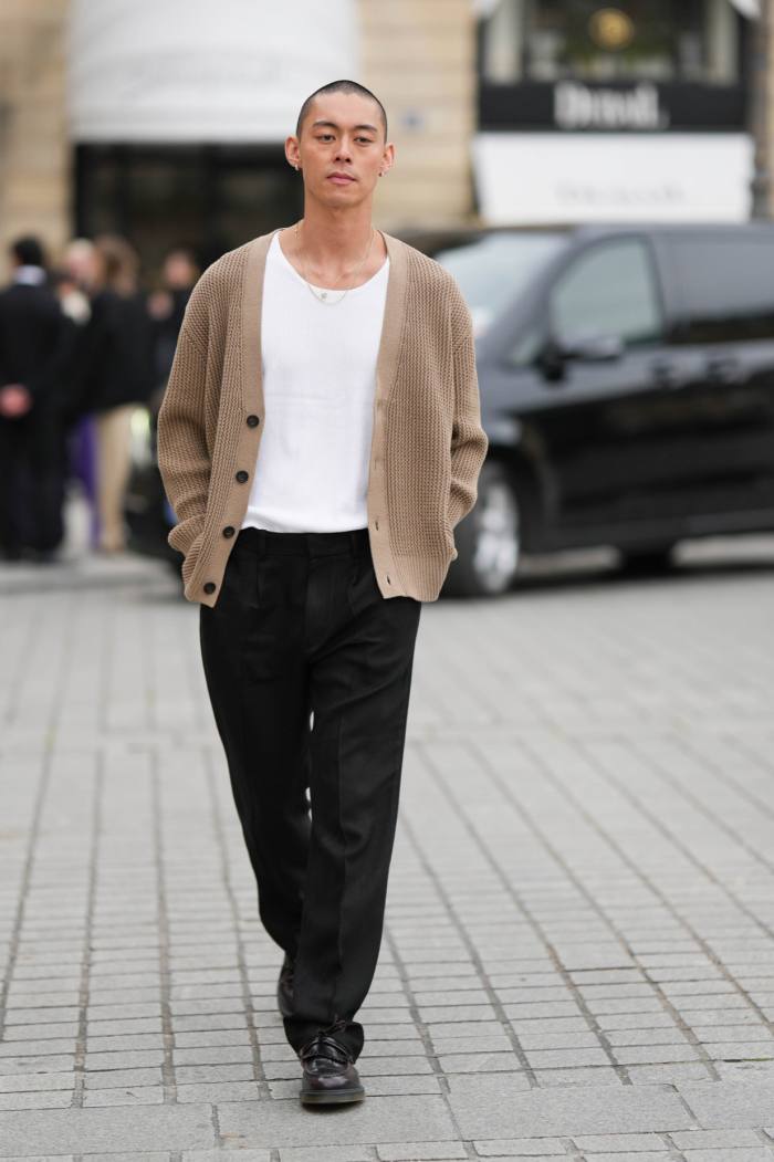 Un oaspete poartă un tricou alb, un cardigan cu nasturi de lână bej, pantaloni negri, pantofi negri cu șireturi din piele lucioasă