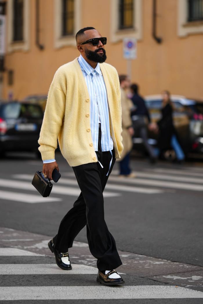 Un oaspete poartă ochelari de soare negri, o cămașă cu imprimeu în dungi albe și albastre, un cardigan din lână galben pal cu nasturi, pantaloni de costum negri, o geantă neagră din piele strălucitoare, mocasini din piele strălucitoare cu imprimeu bicolor alb-negru.