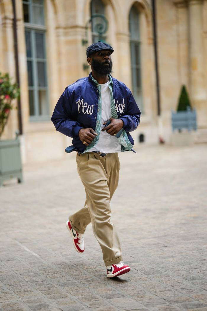 Un oaspete poartă o pălărie din denim bleumarin, cercei cu pandantiv cu perle aurii și albe, un tricou alb, o cămașă cu imprimeu în dungi de culoare verde închis și alb, o nailon bleumarin cu o haină bomber albă brodată cu model New York, inele aurii, bej pantaloni de costum mari, alb și roșu cu model de logo din piele neagră lucioasă, adidasi Air Force One de la Nike