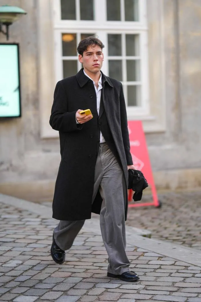 Un oaspete poartă o cămașă albă, un blazer negru, o haină lungă de lână neagră, pantaloni de costum gri, pantofi negri din piele lucioasă