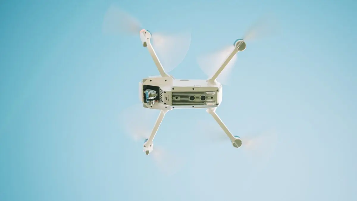 Vedere de jos a unei drone albe (cvadricopter) aflate în zbor, cu elicele în mișcare (efect de blur), pe fundalul unui cer albastru senin