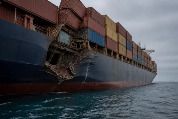 O navă mare de transport maritim aflată pe mare, prezentând o avarie majoră la nivelul carenei și al punții pe partea stângă
