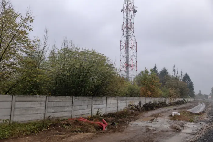 A communication tower stands behind a concrete wall and vegetation near a muddy ground site