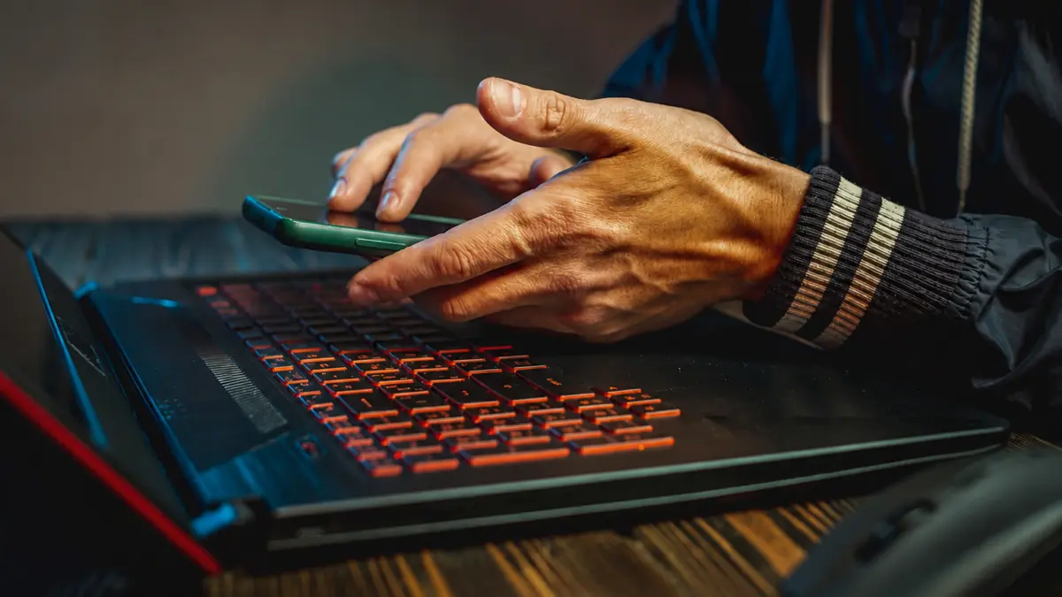 Hands holding a smartphone over a backlit gaming laptop keyboard in a dark setting